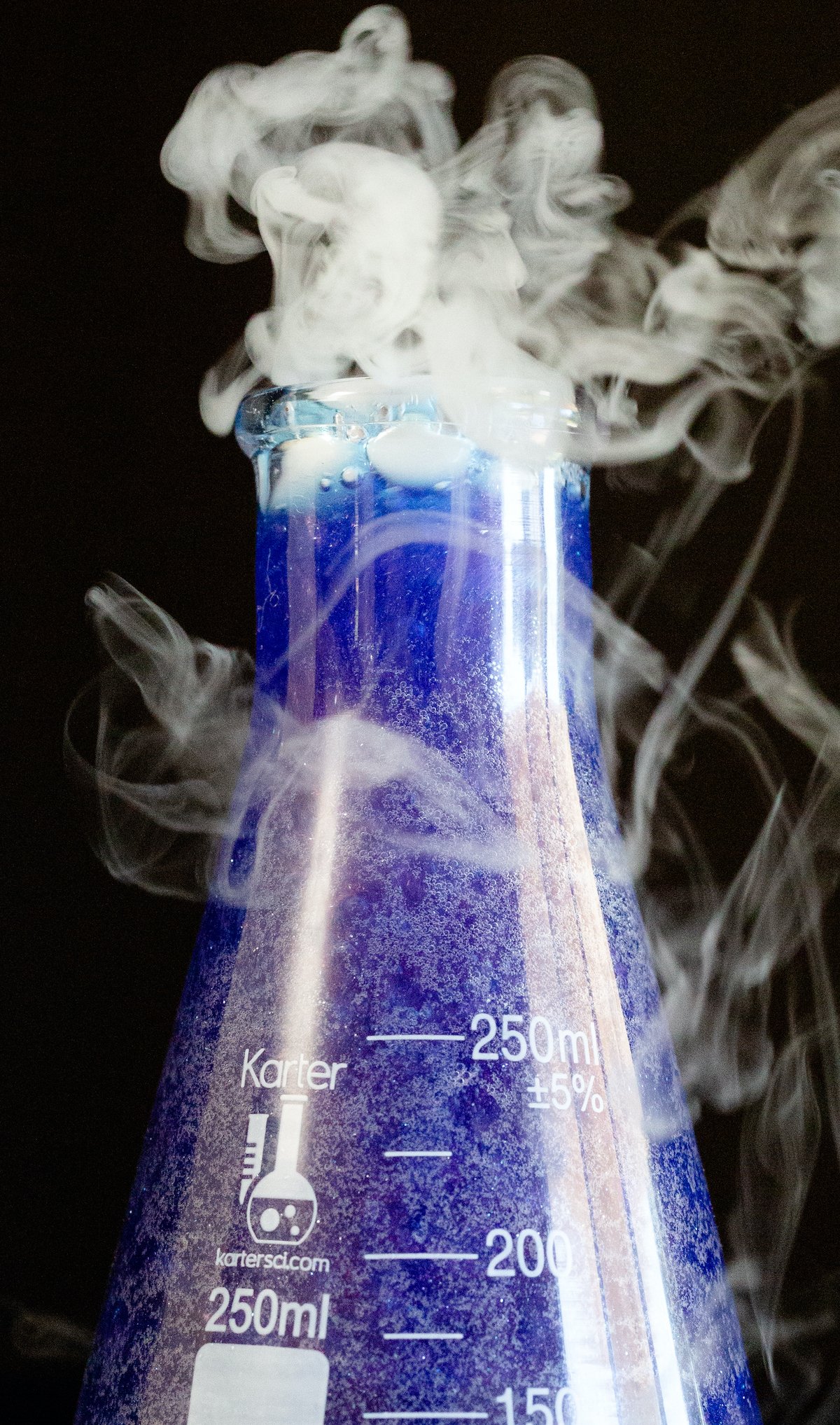 A close up of a purple sparkling witches brew served in a scientific flask cocktail with smoke coming out of the top.