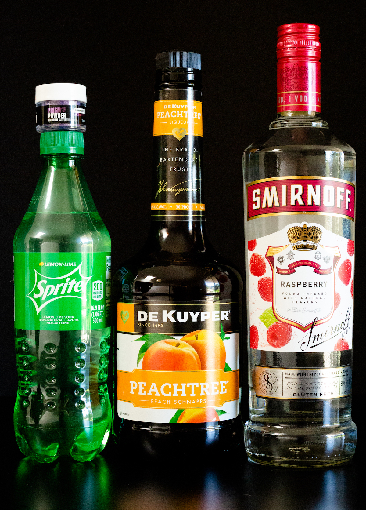 A bottle of Sprite, a small jar of purple prism powder, a bottle of peach liqueur, and a bottle of raspberry vodka sit on a black background.