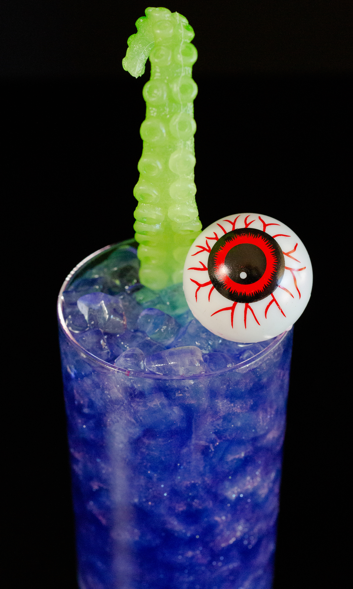 A highball glass is filled with a sparkling purple witches brew and is topped with a plastic eyeball and green sea monster arm.
