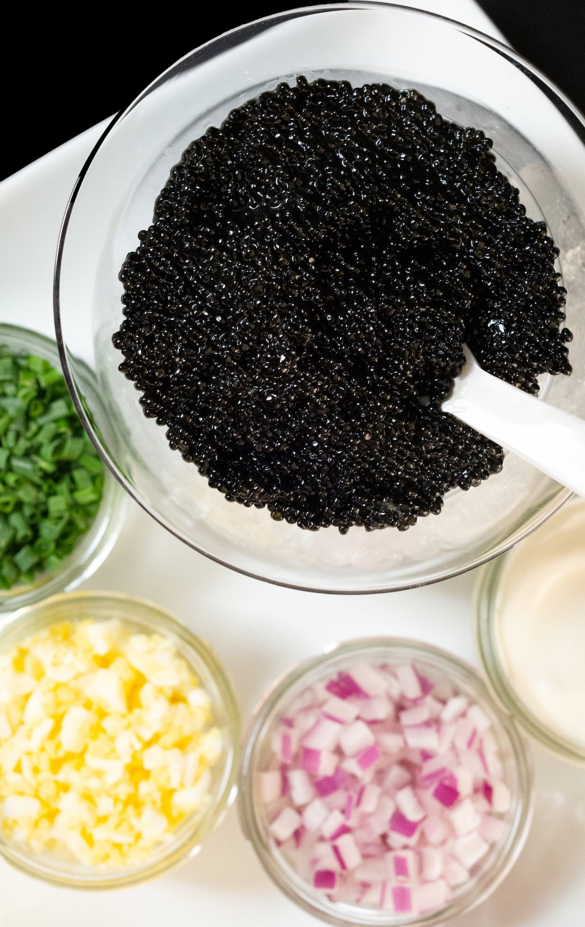 Overhead view of a large bowl of caviar with a pearl spoon. Jars of chopped green onion, red onion, hard boiled egg, and creme fraiche surround it.