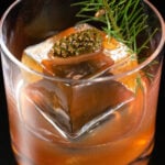 Overhead view of a lowball glass on a black background. It is filled half way with a reddish brown Christmas old fashioned liquid. It also has a large clear block of ice and a mini pine cone and small clipping from a Christmas tree as a garnish.