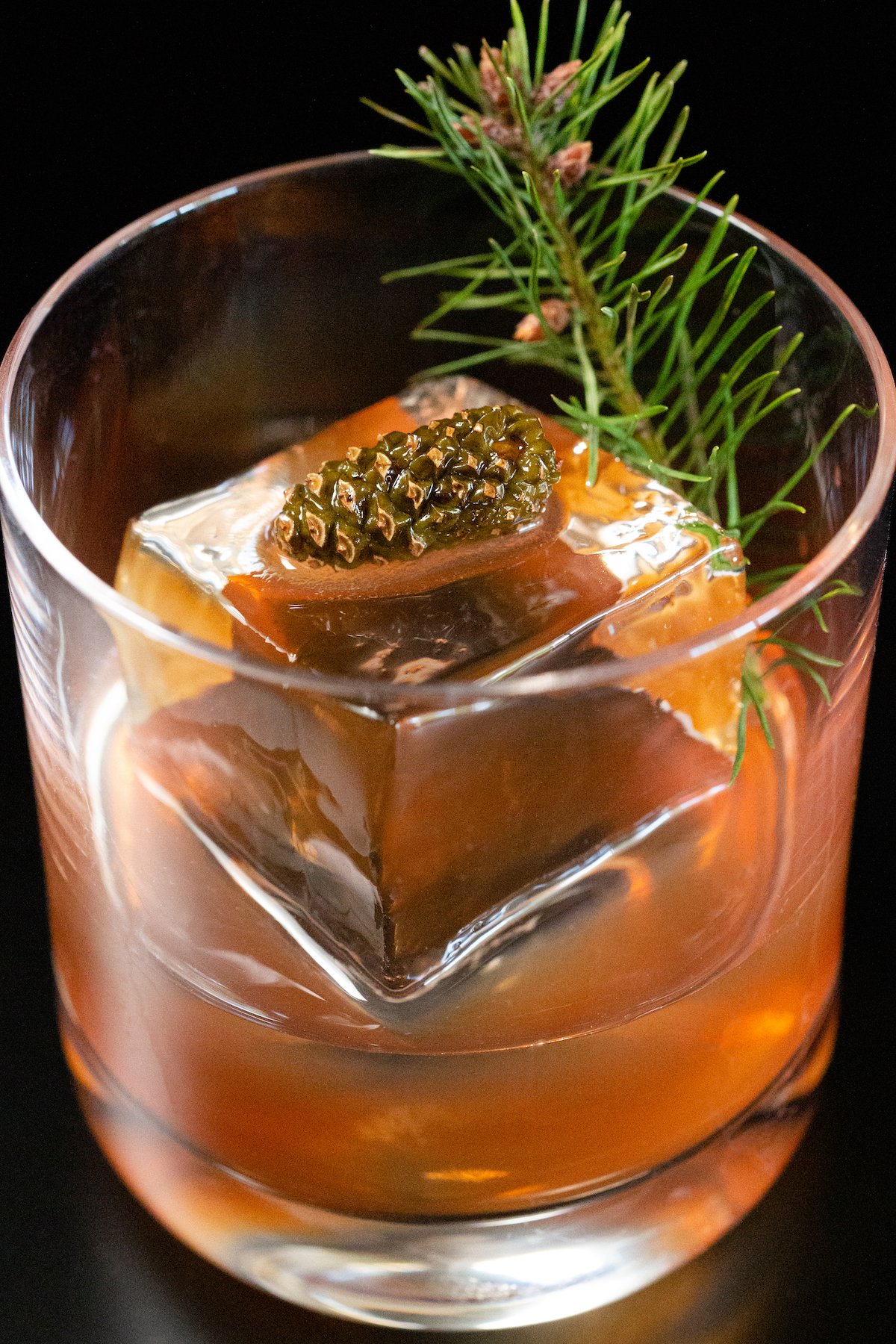Overhead view of a lowball glass on a black background. It is filled half way with a reddish brown Christmas old fashioned liquid. It also has a large clear block of ice and a mini pine cone and small clipping from a Christmas tree as a garnish.