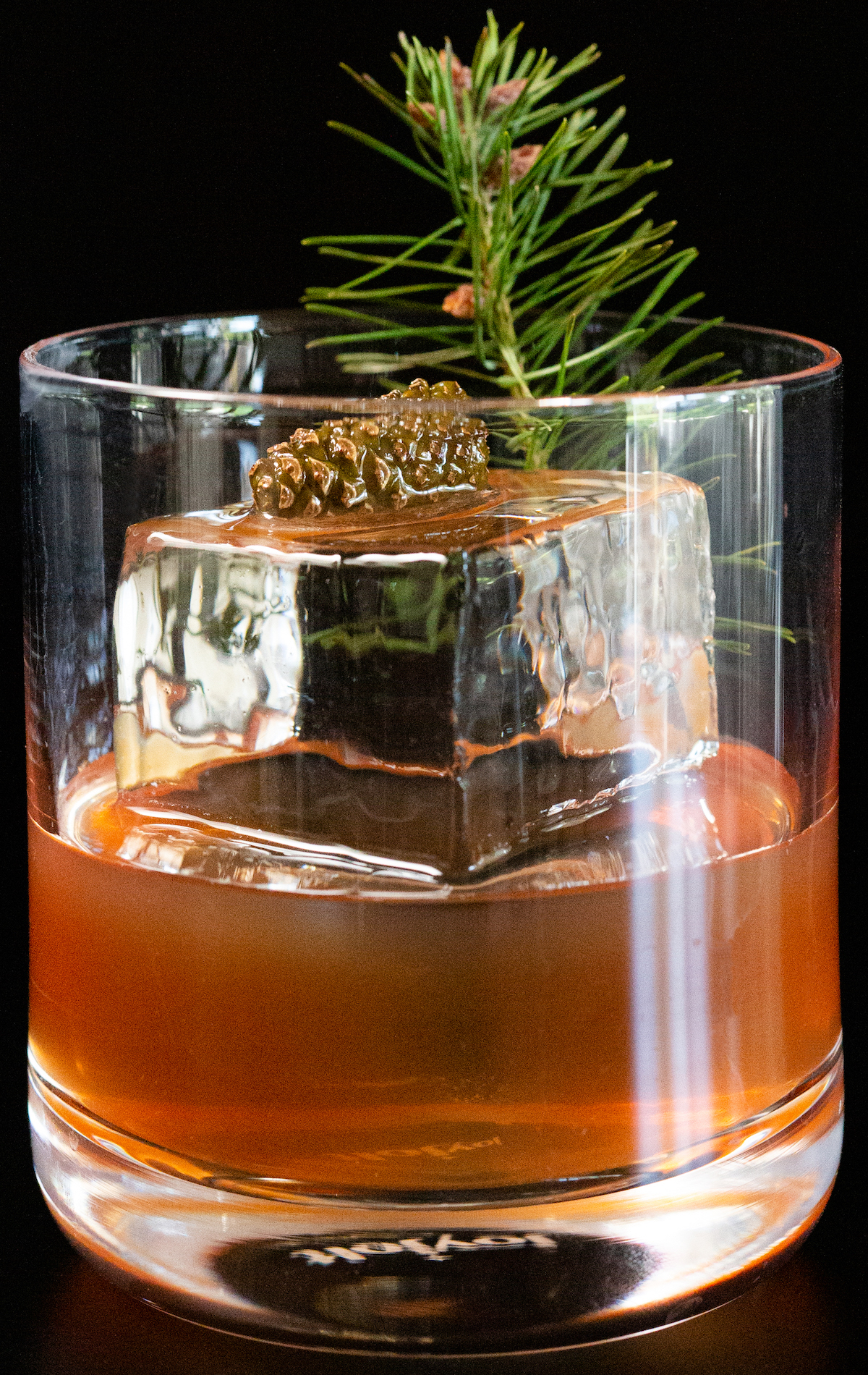 A lowball glass on a black background is filled halfway with a reddish brown Christmas old fashioned. There is a large block of clear ice and a small pine cone and clipping from a Christmas as garnishes.