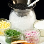 A glass cone is filled with caviar and sits on top of a glass bowl filled with ice. Around it are jars of chopped boiled egg, green onion, red onion, and creme fraiche as well as blinis.