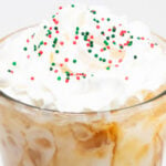 Close up of a pint glass that's filled with coffee and almond milk then topped with whipped cream and red, white, and green Christmas sprinkles.
