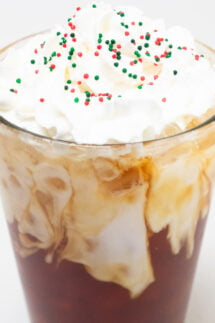 Close up of a pint glass that's filled with coffee and almond milk then topped with whipped cream and red, white, and green Christmas sprinkles.