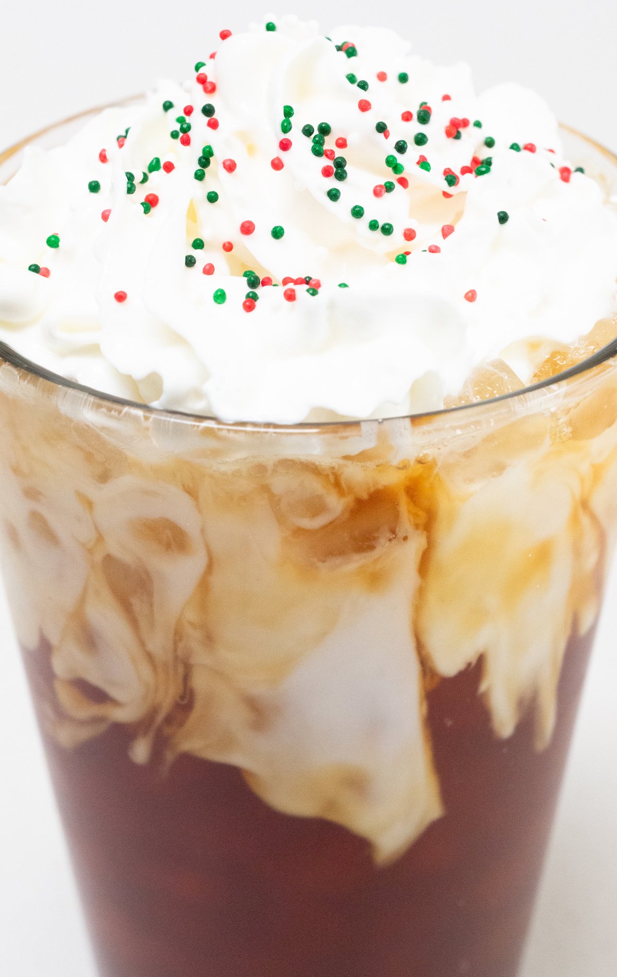 Close up of a pint glass that's filled with coffee and almond milk then topped with whipped cream and red, white, and green Christmas sprinkles.