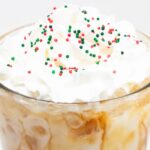 Close up of a pint glass that's filled with coffee and almond milk then topped with whipped cream and red, white, and green Christmas sprinkles.