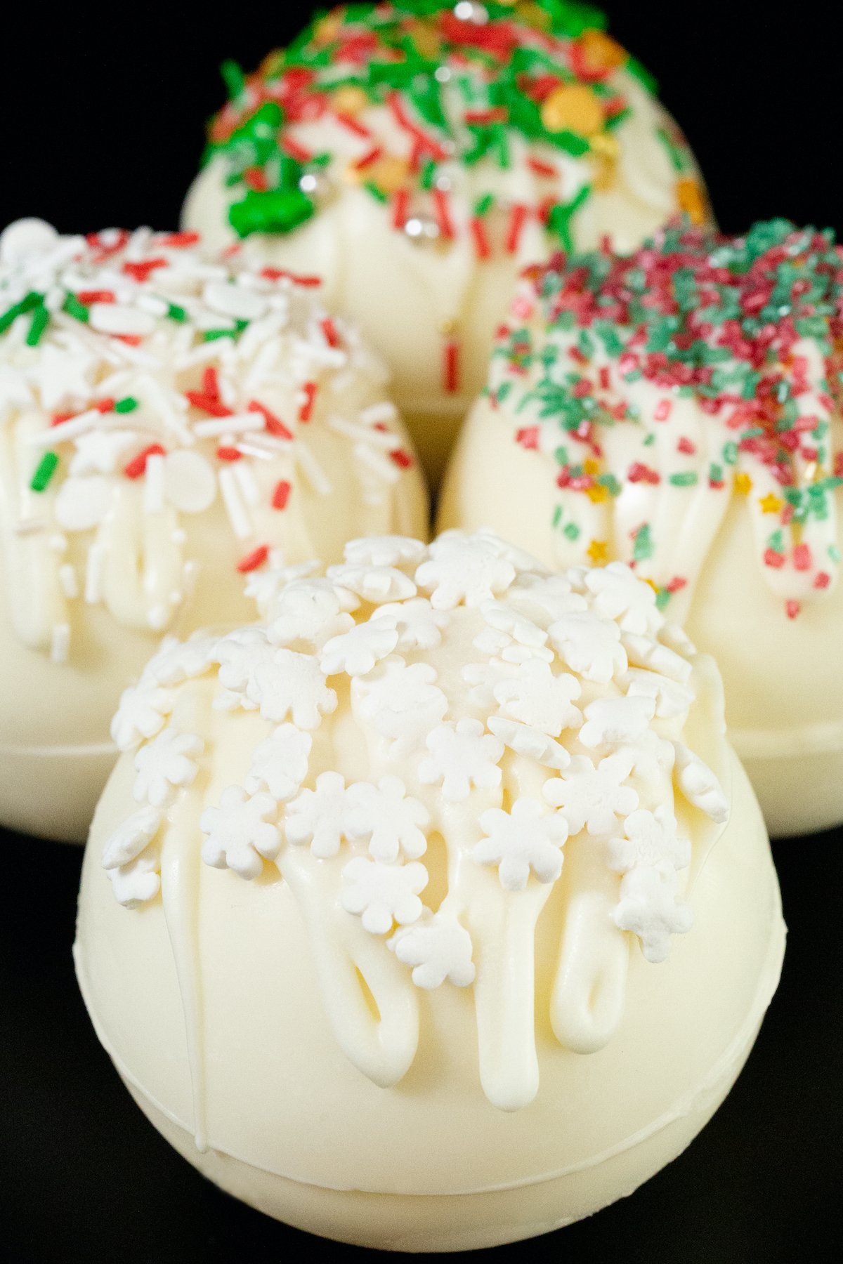 4 white hot chocolate bombs sit on a black background. They are adorned with drizzled white chocolate at various Christmas colored sprinkles.