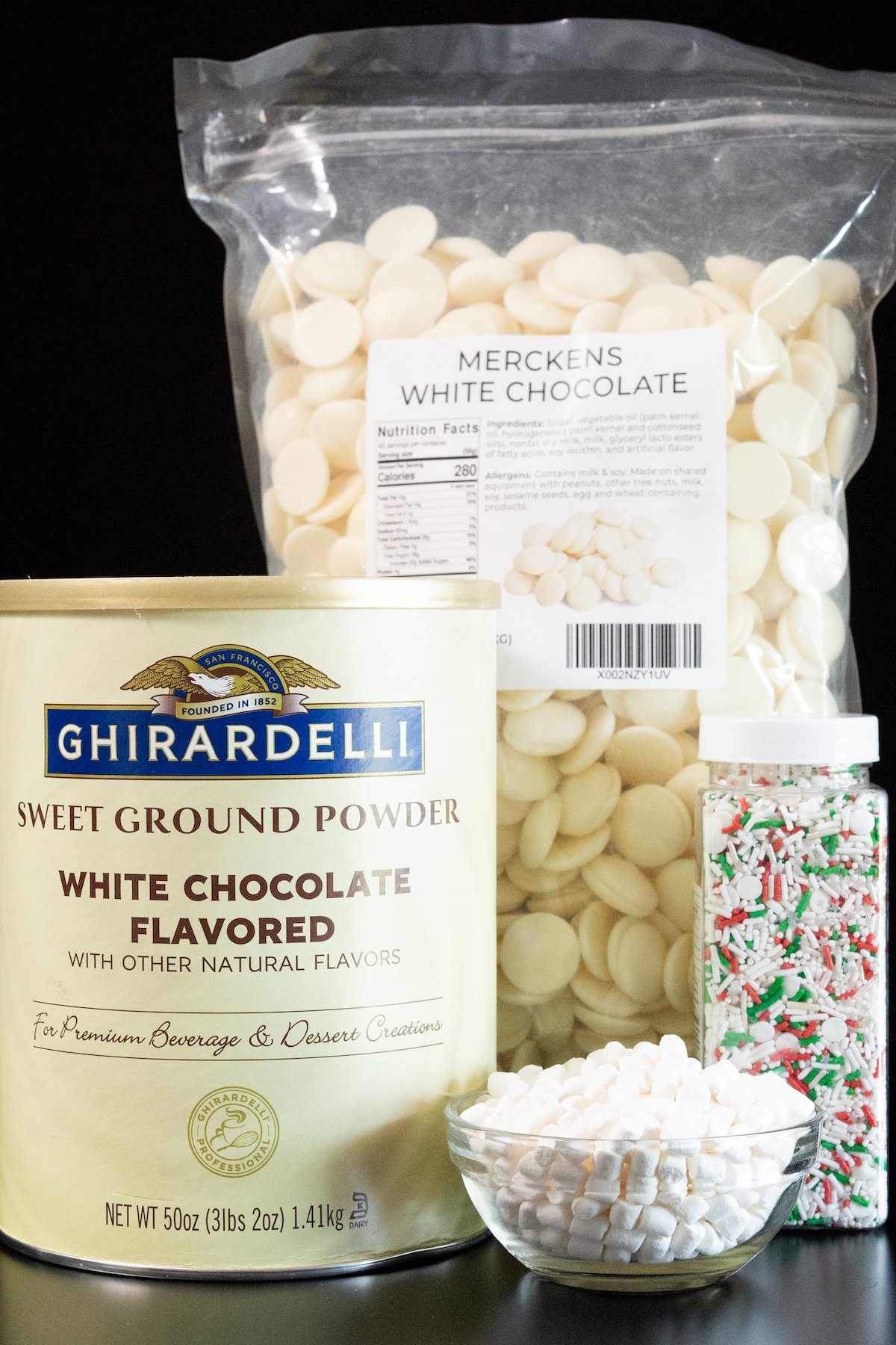 All the ingredients to make white hot chocolate bombs sit on a black background - a tub of Ghirardelli white chocolate powder, dehydrated marshmallows, white chocolate melts, and sprinkles.