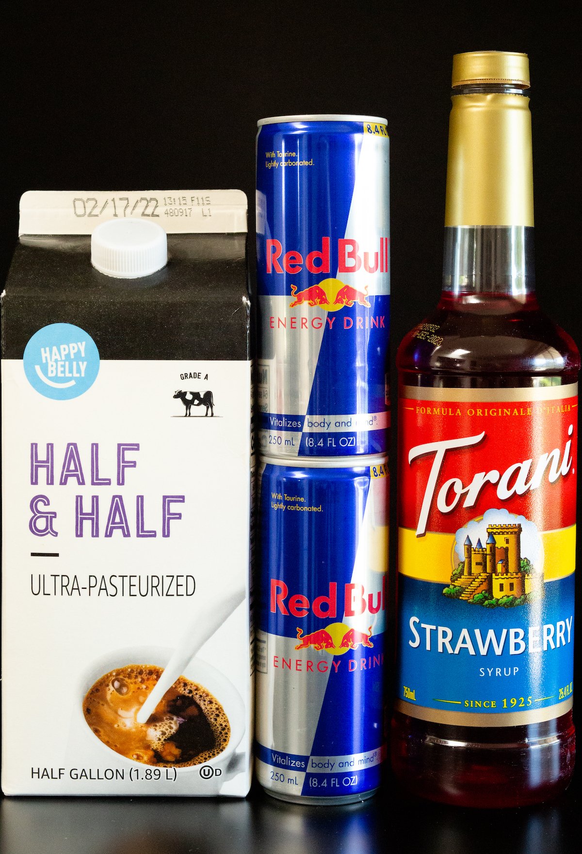 A carton of half and half, two cans of red bull, and a strawberry syrup bottle on a black background.