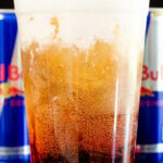 A large plastic glass filled with Red Bull Italian Soda. It has distinct layers of raspberry syrup, red bull soda, and cream on top.