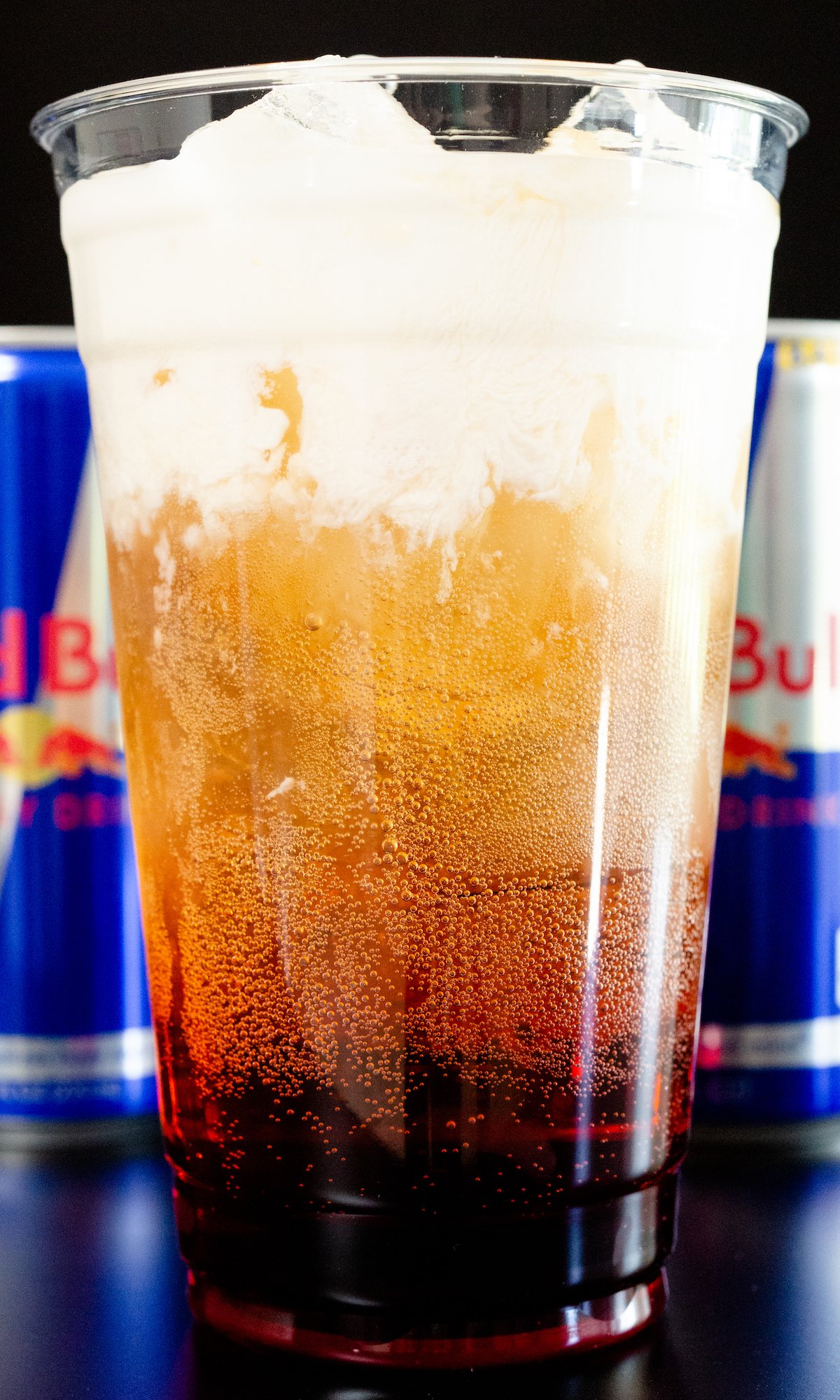 A large plastic glass filled with Red Bull Italian Soda. It has distinct layers of raspberry syrup, red bull soda, and cream on top.