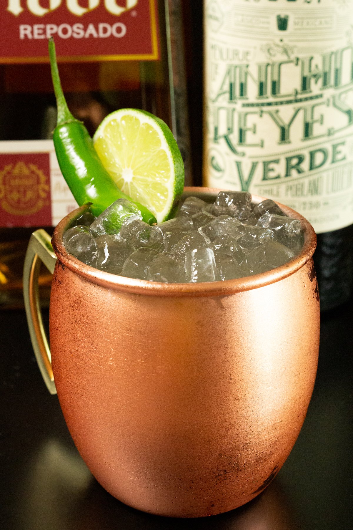 A copper mug filled with pebble ice and Mexican Mule in front of a tequila and chile liqueur bottle.