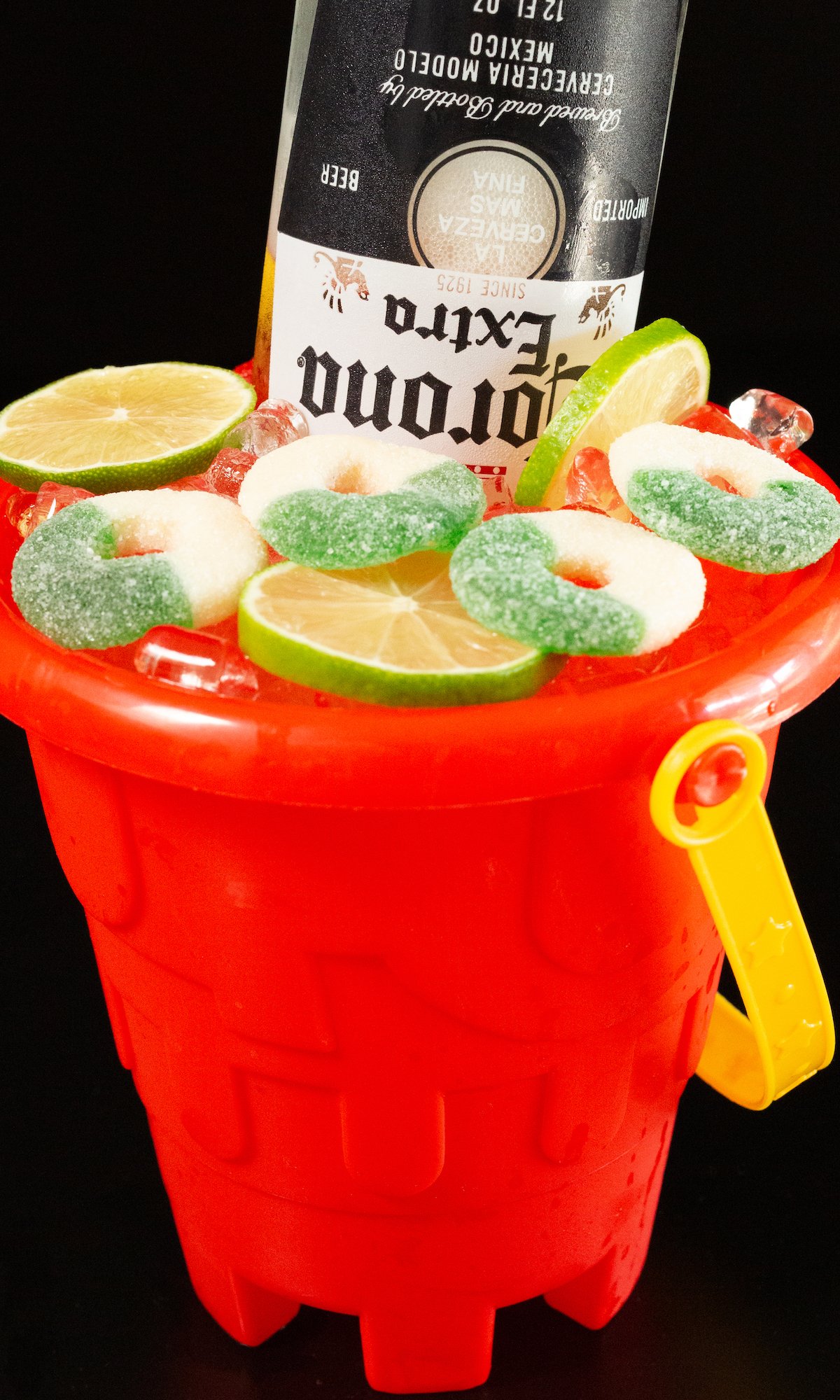 A red sand bucket filled with a tequila cocktail that's topped with sliced limes, green apple gummy rings, and a Mexican beer.