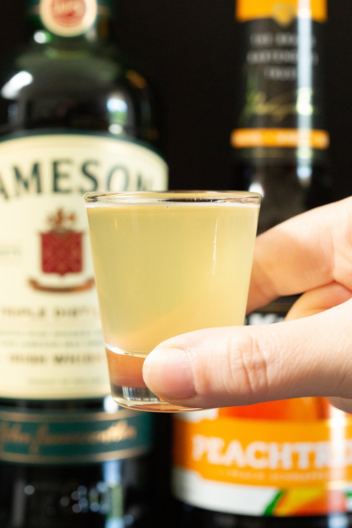 A hand holds up a clear shot glass filled with a green tea shot.