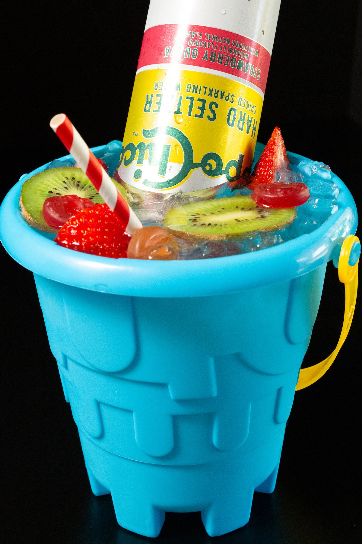 A sand bucket is filled with a cocktail that's topped with sliced kiwis, strawberries, lifesavers, and a hard seltzer.