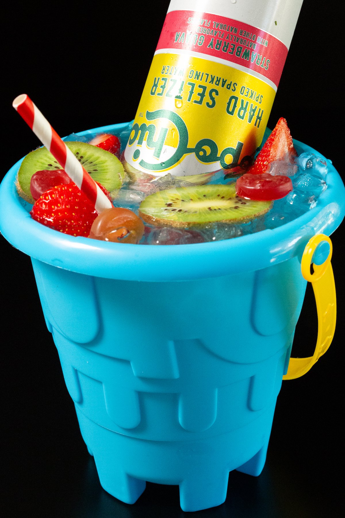 A blue sand bucket filled with a cocktail that's topped with sliced strawberries, kiwis, and gummy lifesavers.
