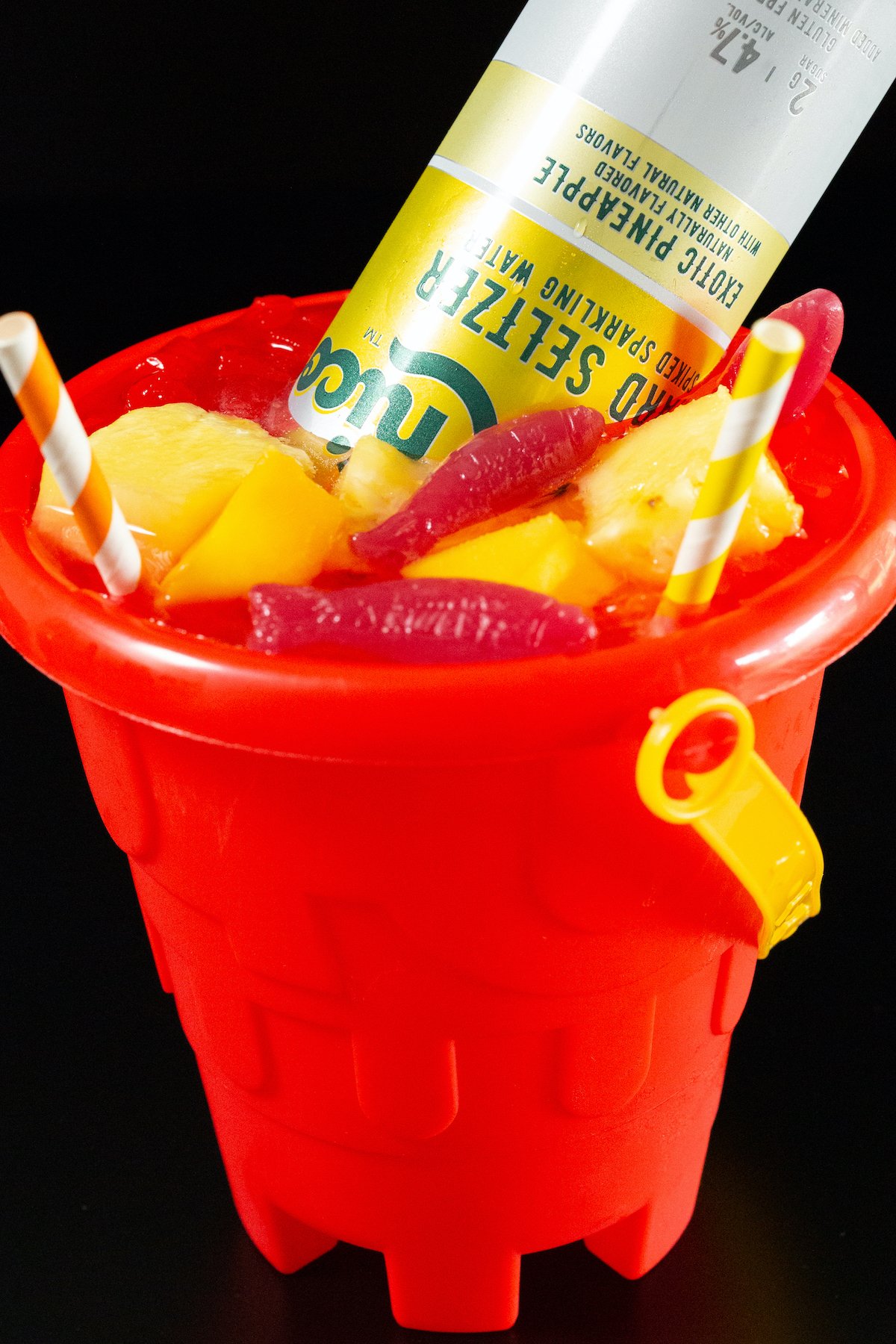 A red sand bucket filled with a cocktail that's topped with fresh fruit, swedish fish candy, and a hard seltzer.