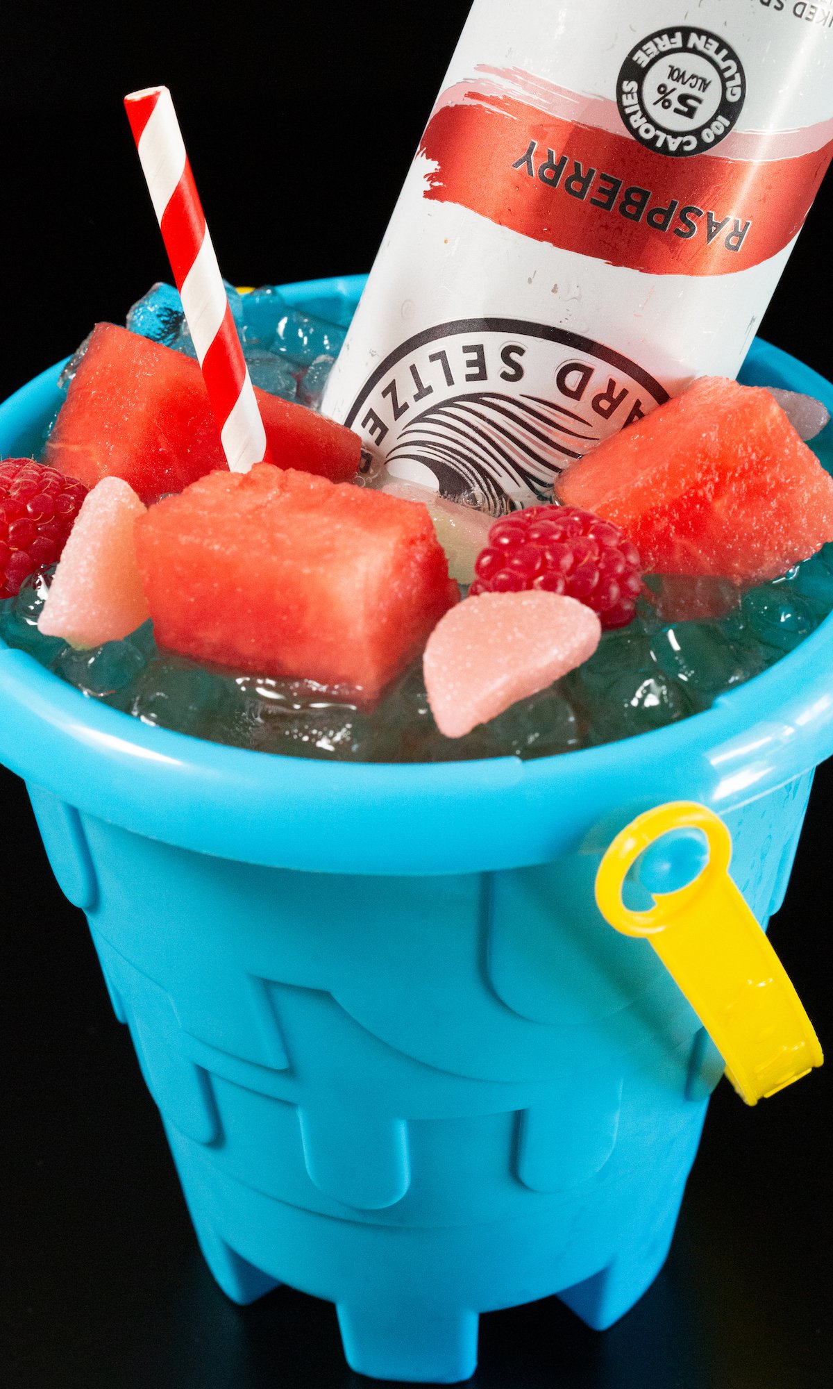 A sand bucket filled with a cocktail that's topped with sliced watermelon, raspberries, watermelon candy, and White Claw.