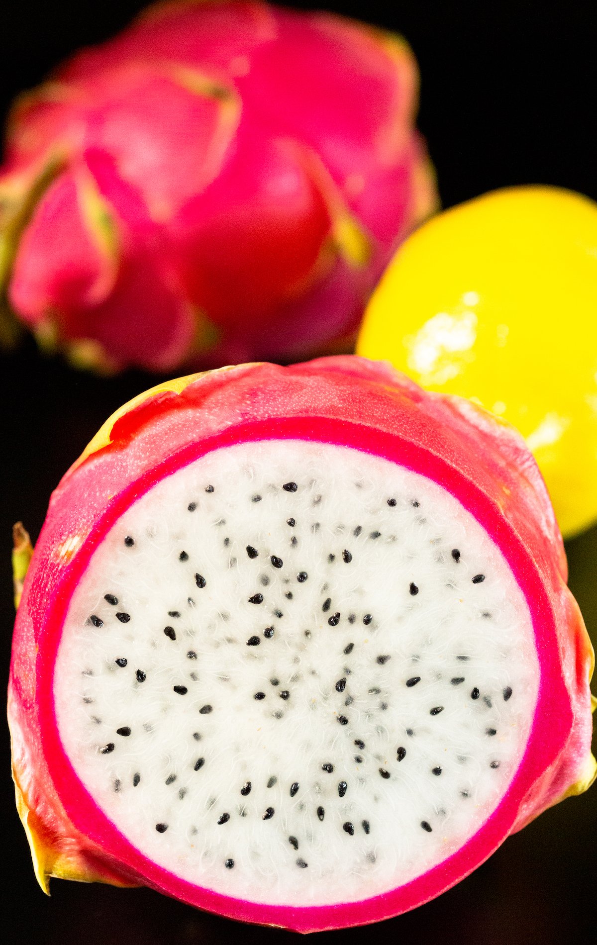A dragonfruit sliced in half to show the white flesh inside sits in front if a lemon and an uncut dragonfruit. 