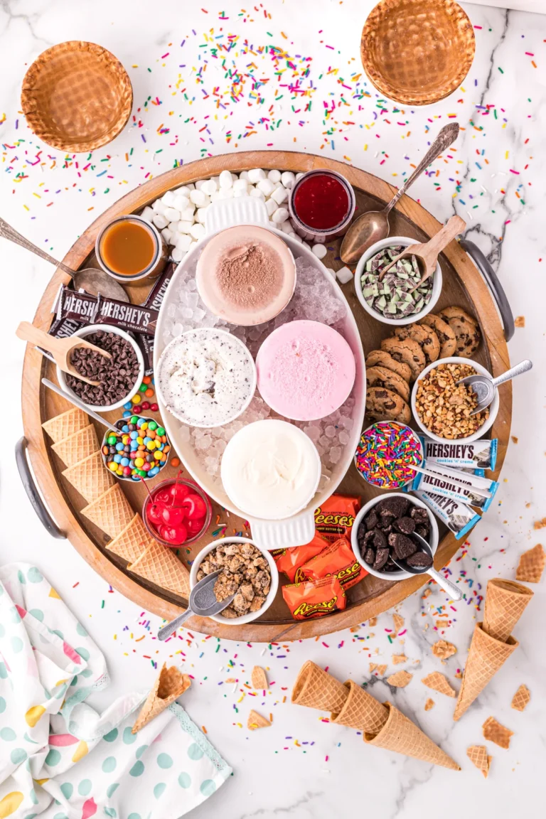 Overhead view of a deep round wooden board filled with 4 different pints of ice cream, waffle cones, and assorted toppings.