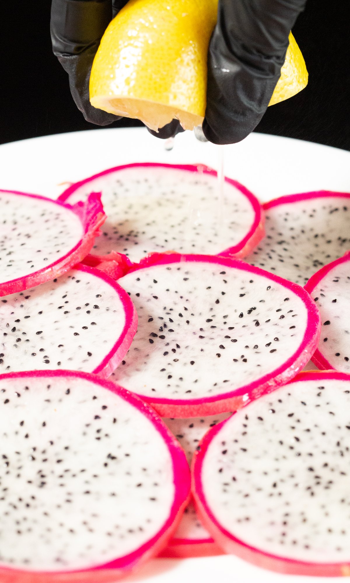 Dragonfruit slices in a single layer on a white plate. A hand squeezes fresh lemon juice over the top.