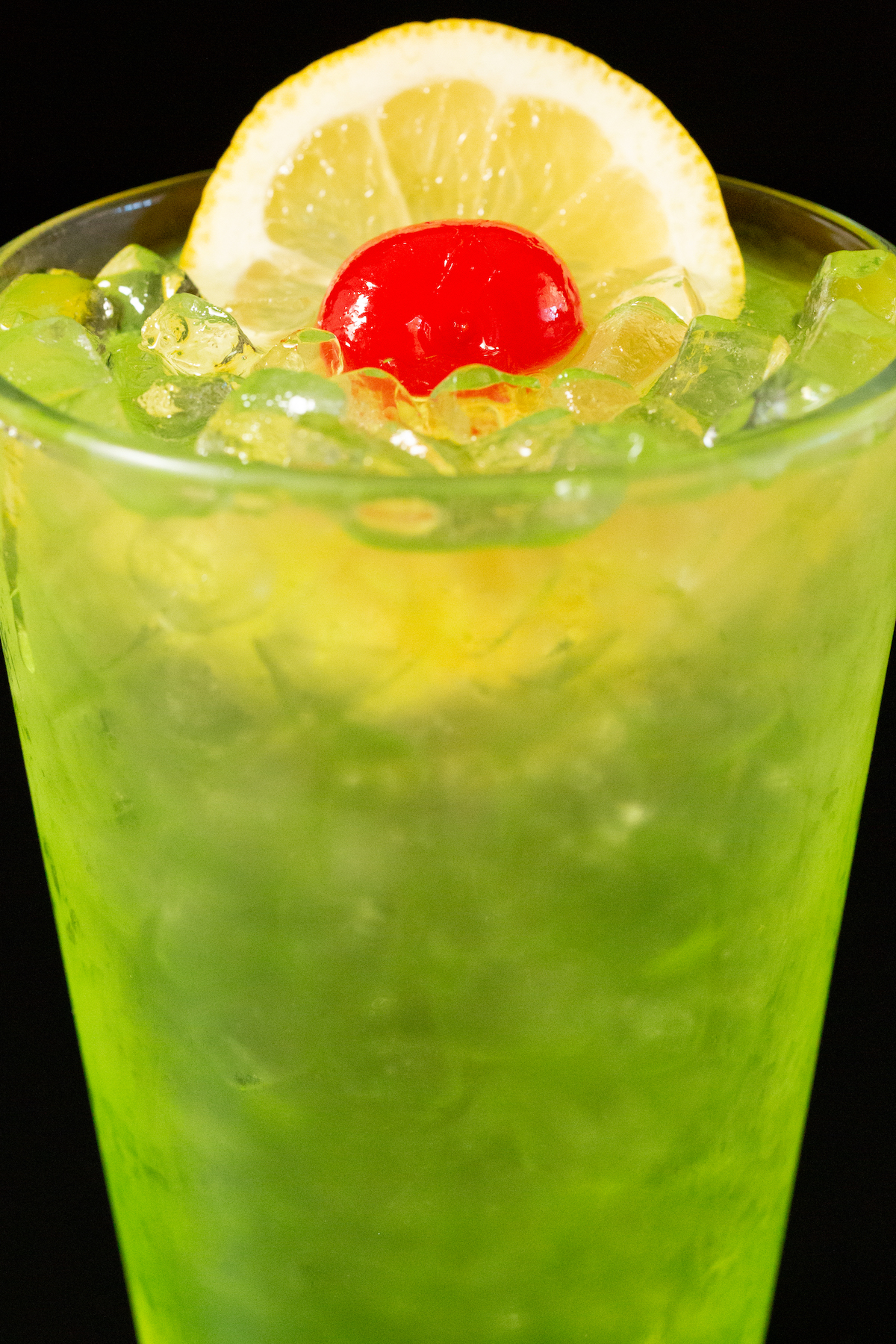 Close up of a pint glass filled with a neon green Tokyo Iced Tea Cocktail that's topped with a lime wheel and maraschino cherry.