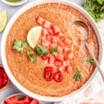 A white soup bowl filled with chipotle watermelon gazpacho. It is garnished with cilantro, a lime wedge, red pepper slices, and diced tomato.