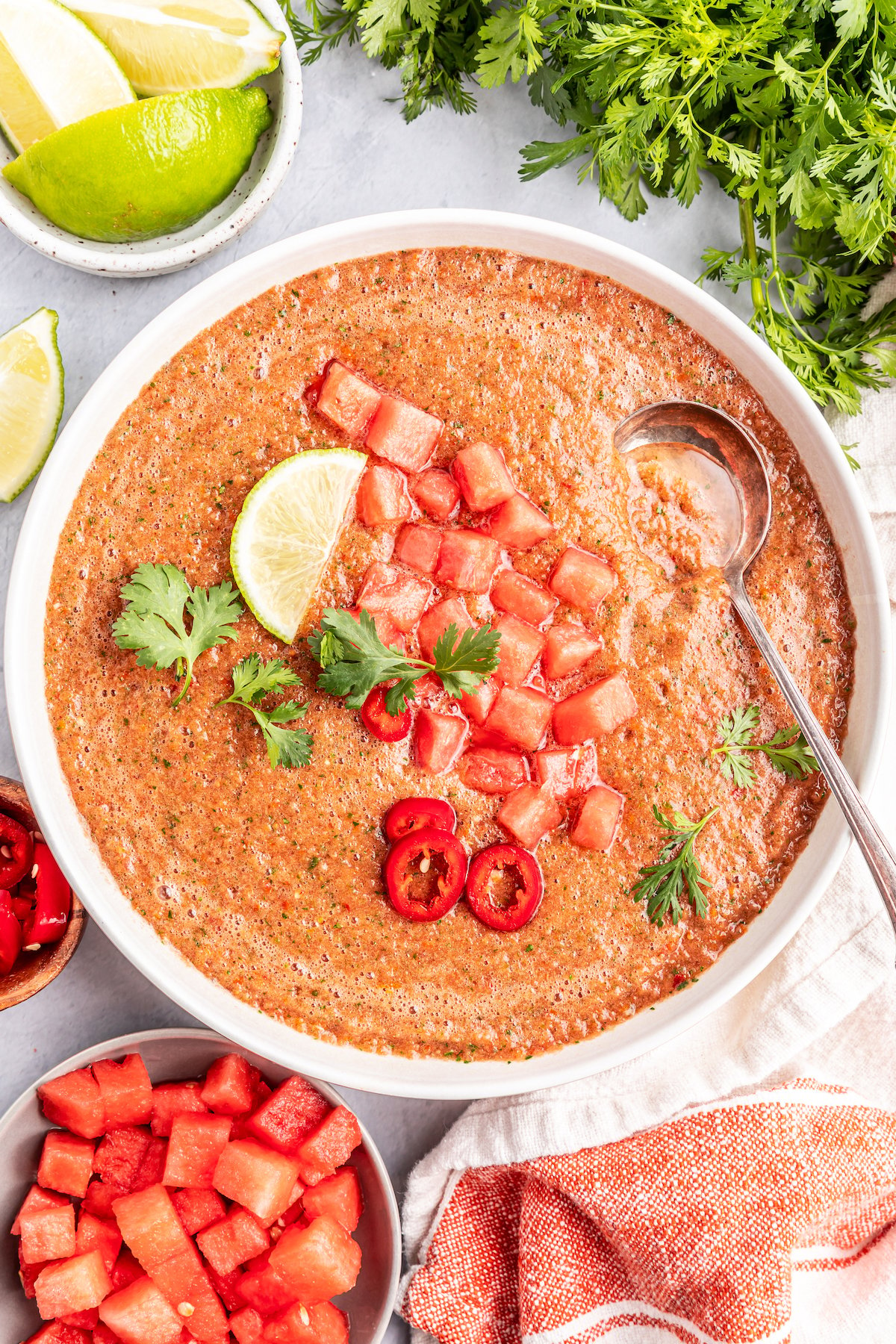 A white soup bowl filled with chipotle watermelon gazpacho. It is garnished with cilantro, a lime wedge, red pepper slices, and diced tomato.