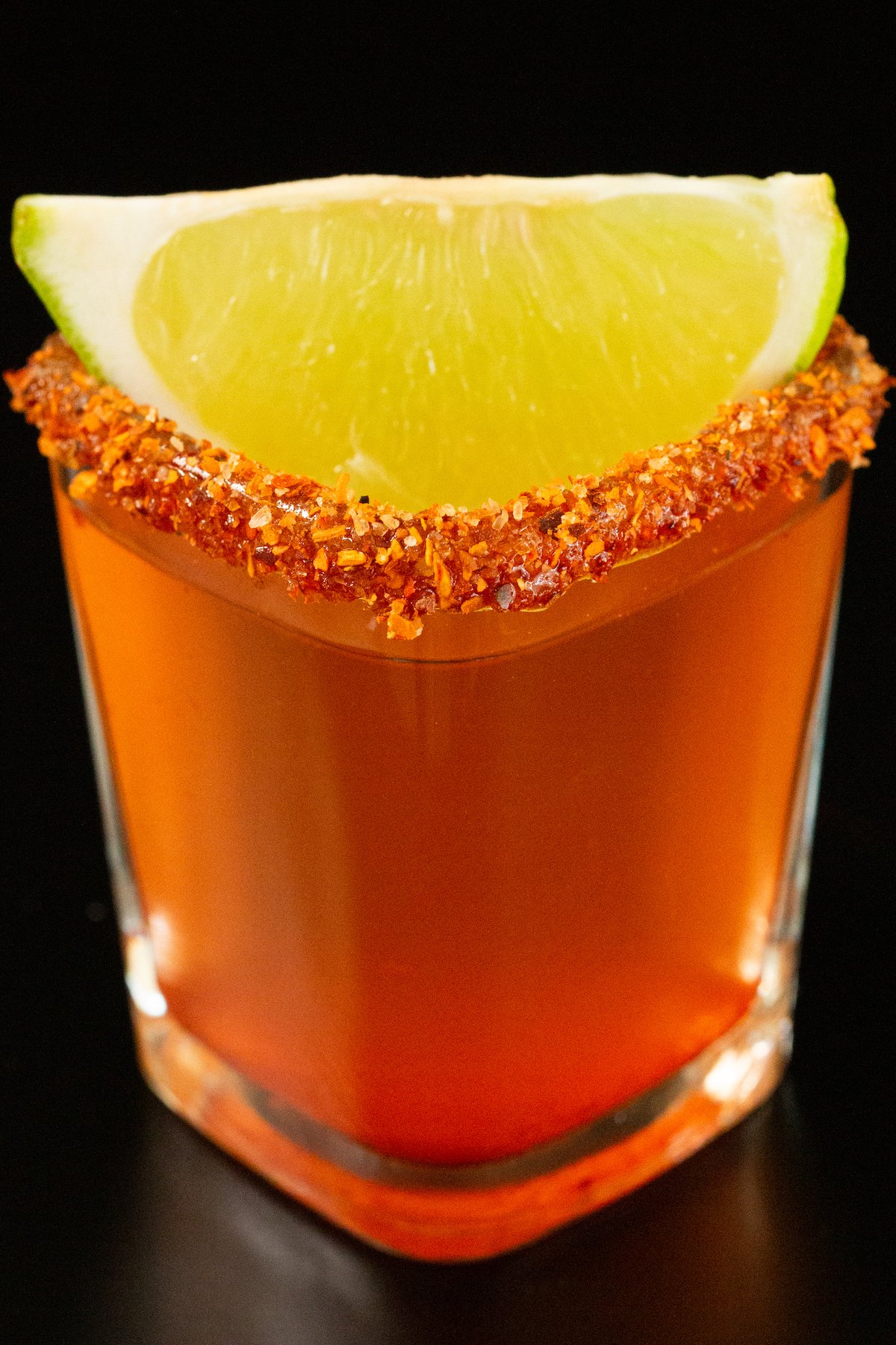 A single square shot glass has been filled with a pinkish-red Mexican candy shot. It is garnished with a chili powder rim and a lime wedge.