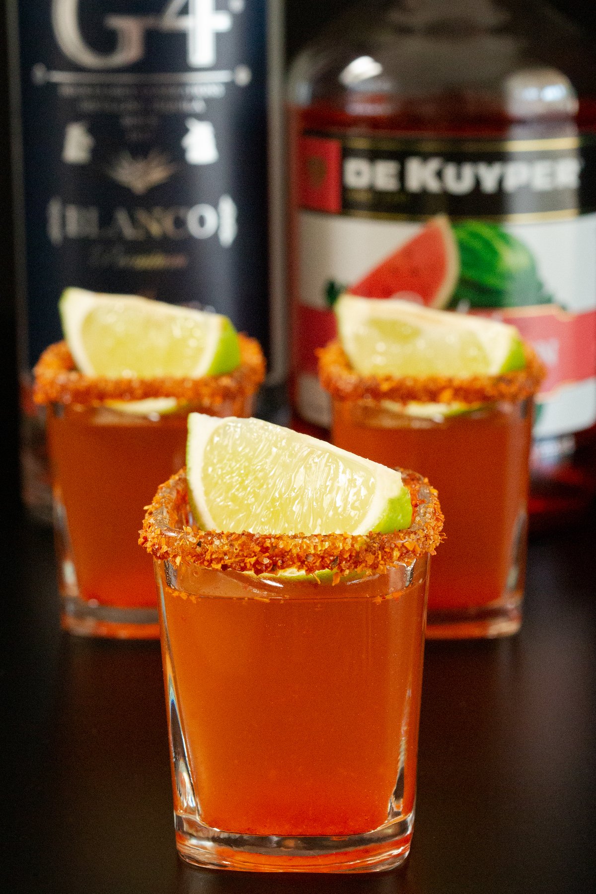 Three square Mexican Candy Shots in front of a bottle of tequila and watermelon schnapps.