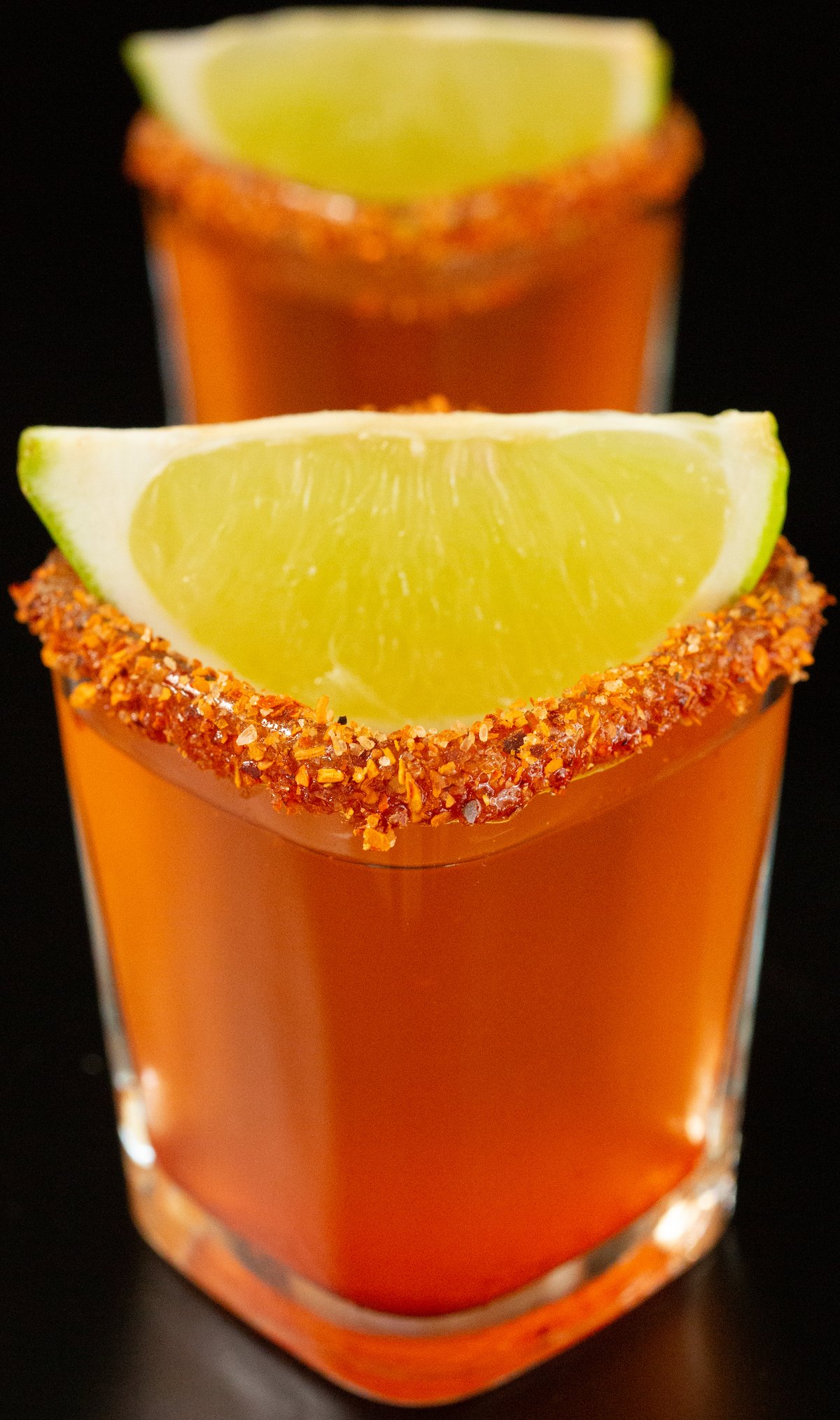 Two square shot glasses filled with a pinkish-red Mexican Candy shot. There is a lime wedge garnish and a chili powder coated rim. 