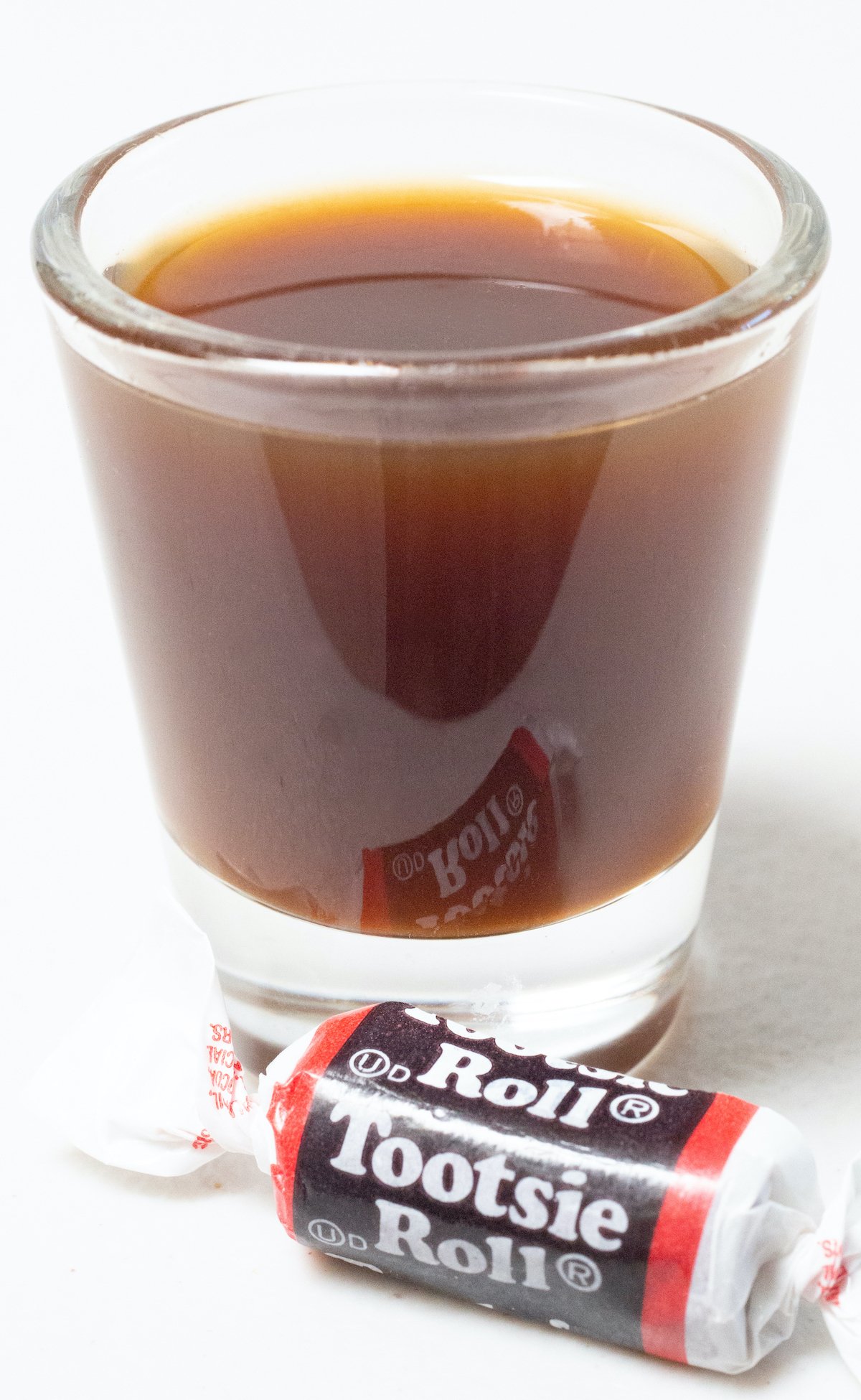 A single shot glass on a white background filled with a brown tootsie roll shot. A piece of candy sits in front.