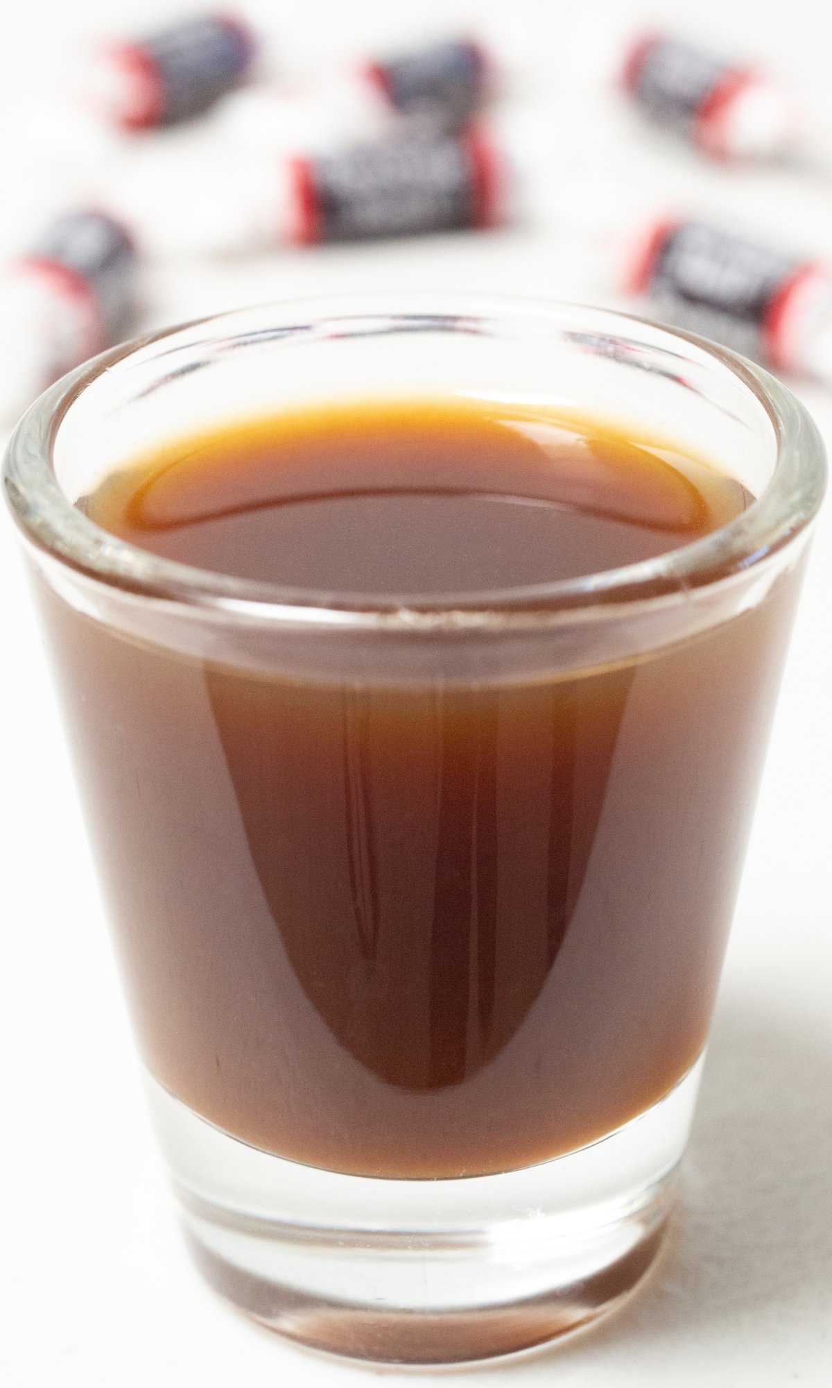 A single shot glass on a white background filled with a brown tootsie roll shot. A piece of candy sits in front.