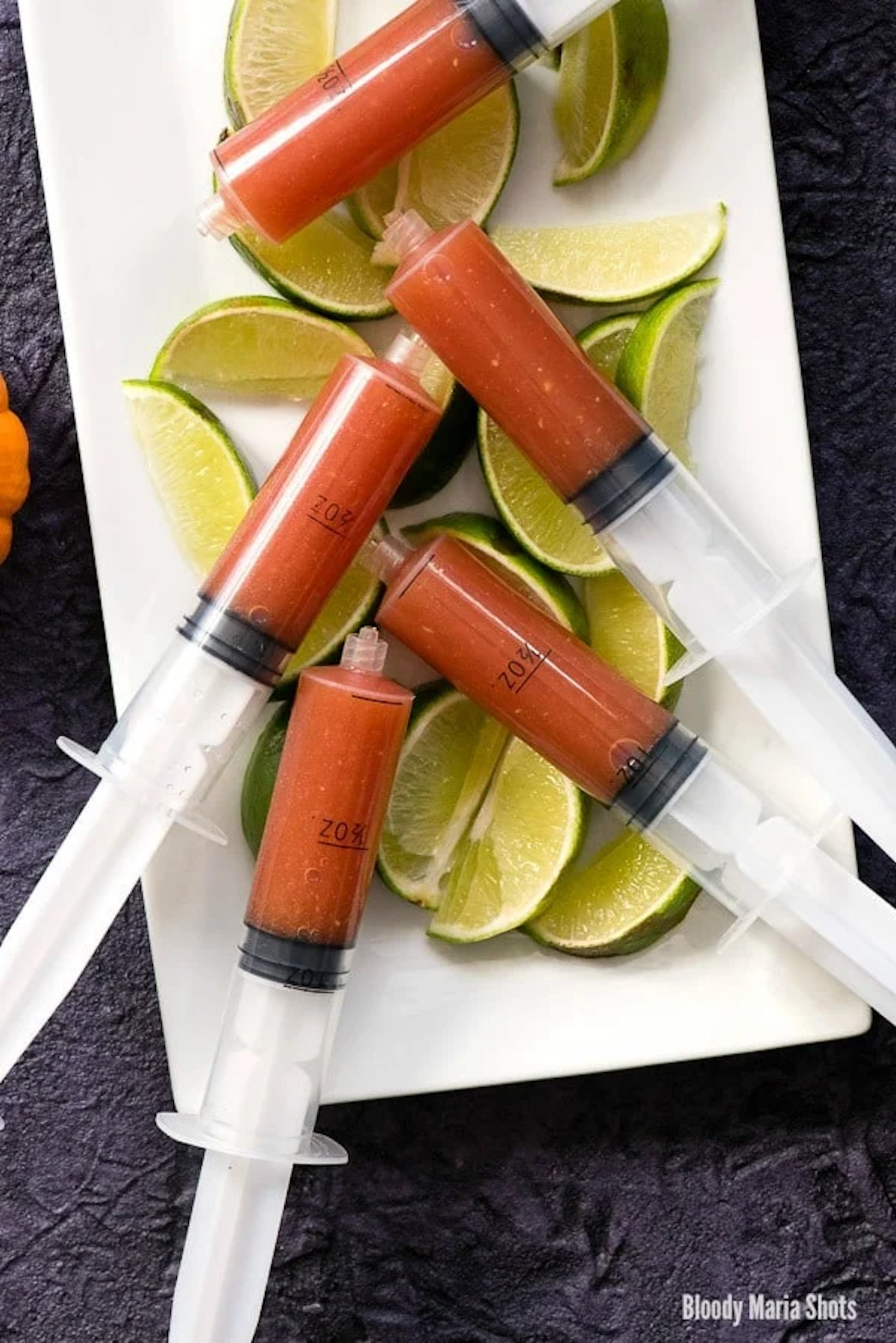 A white serving plate is filled with lime slices that have plastic syringes filled with a red bloody Maria mixture.