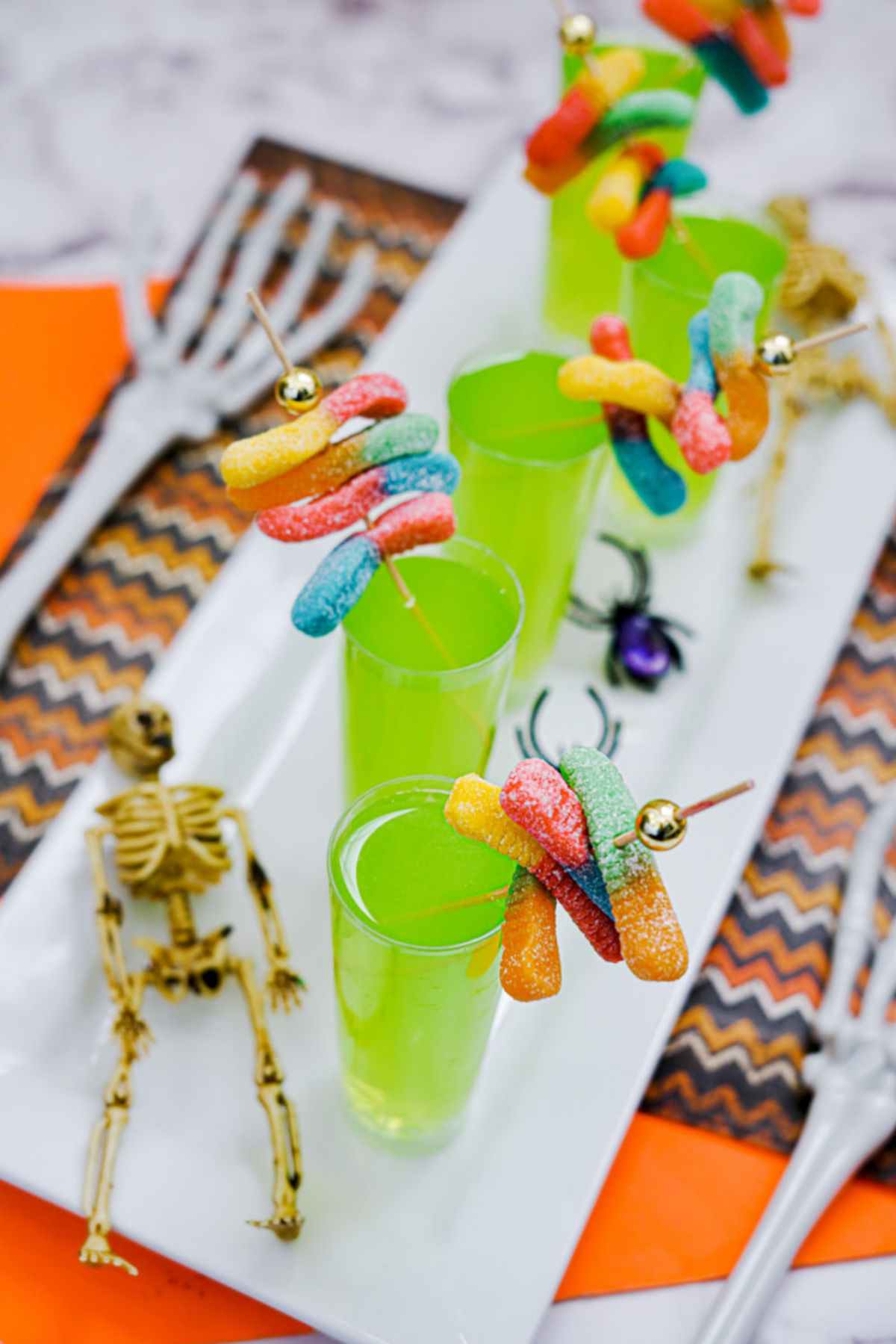 Tall shot glasses are filled with a bright neon green liquid and garnished with sour gummy worms on cocktail skewers.