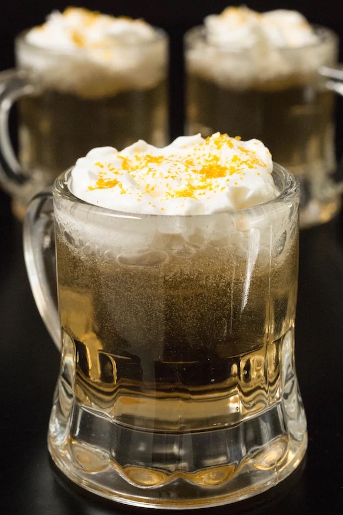 A shotglass sized beer mug is filled with a light brown liquid and topped with whipped cream and gold flakes.
