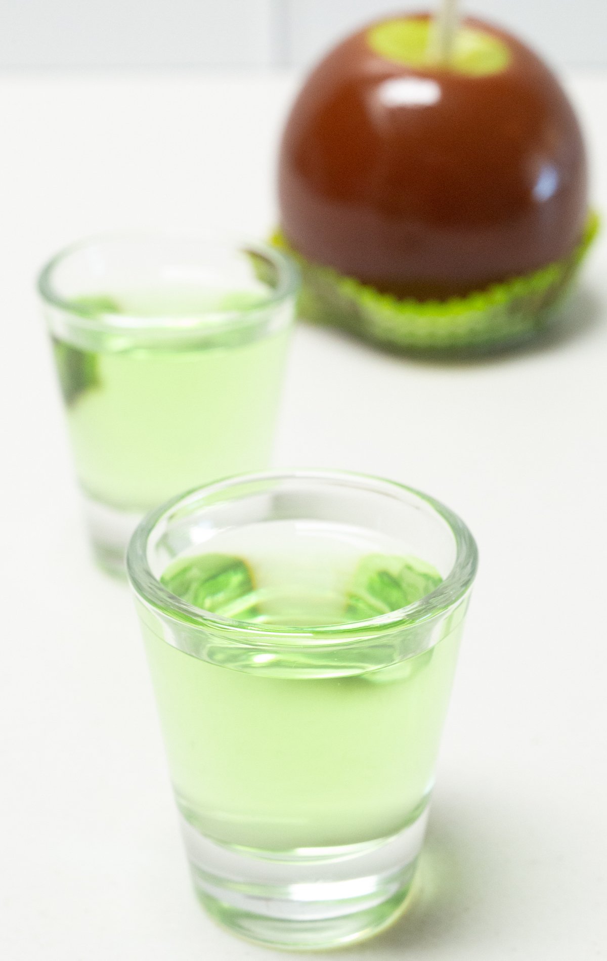 Two shot glasses filled with green caramel apple shots on a white background.