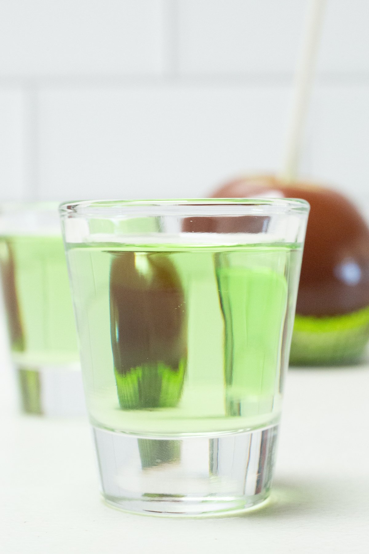 A shot glass filled with a green caramel apple shot is in focus in the foreground, a caramel apple is out of focus in the background.