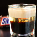 A square shot glass is filled with a snickers shot. You can see the three distinct liqueurs layered.