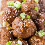 Dozens of asian meatballs covered in brown sauce, sesame seeds, and sliced green onions.