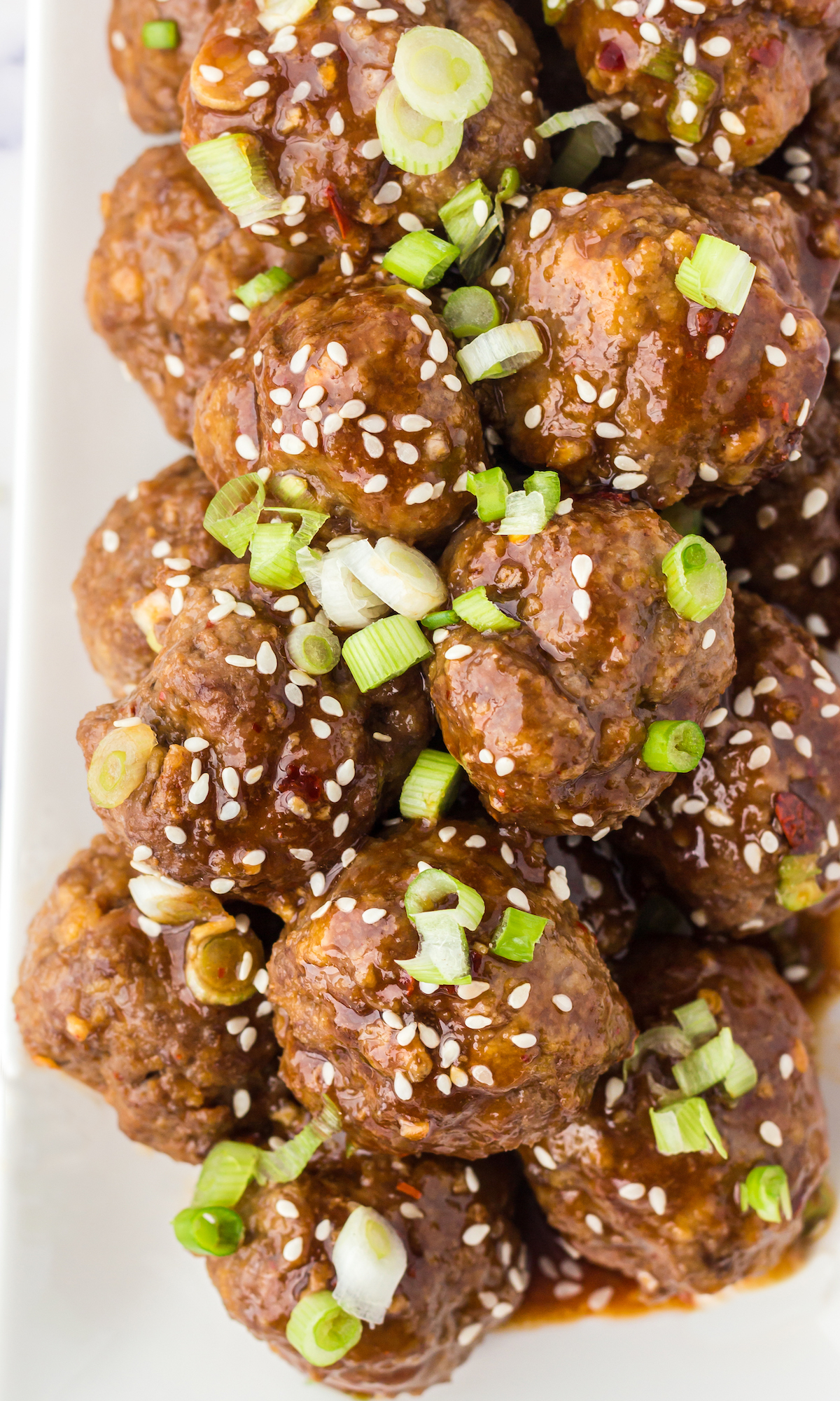 Overhead view of a few dozen Asian meatballs in brown sauce on a white plate. Garnished with sliced green onions and sesame seeds.