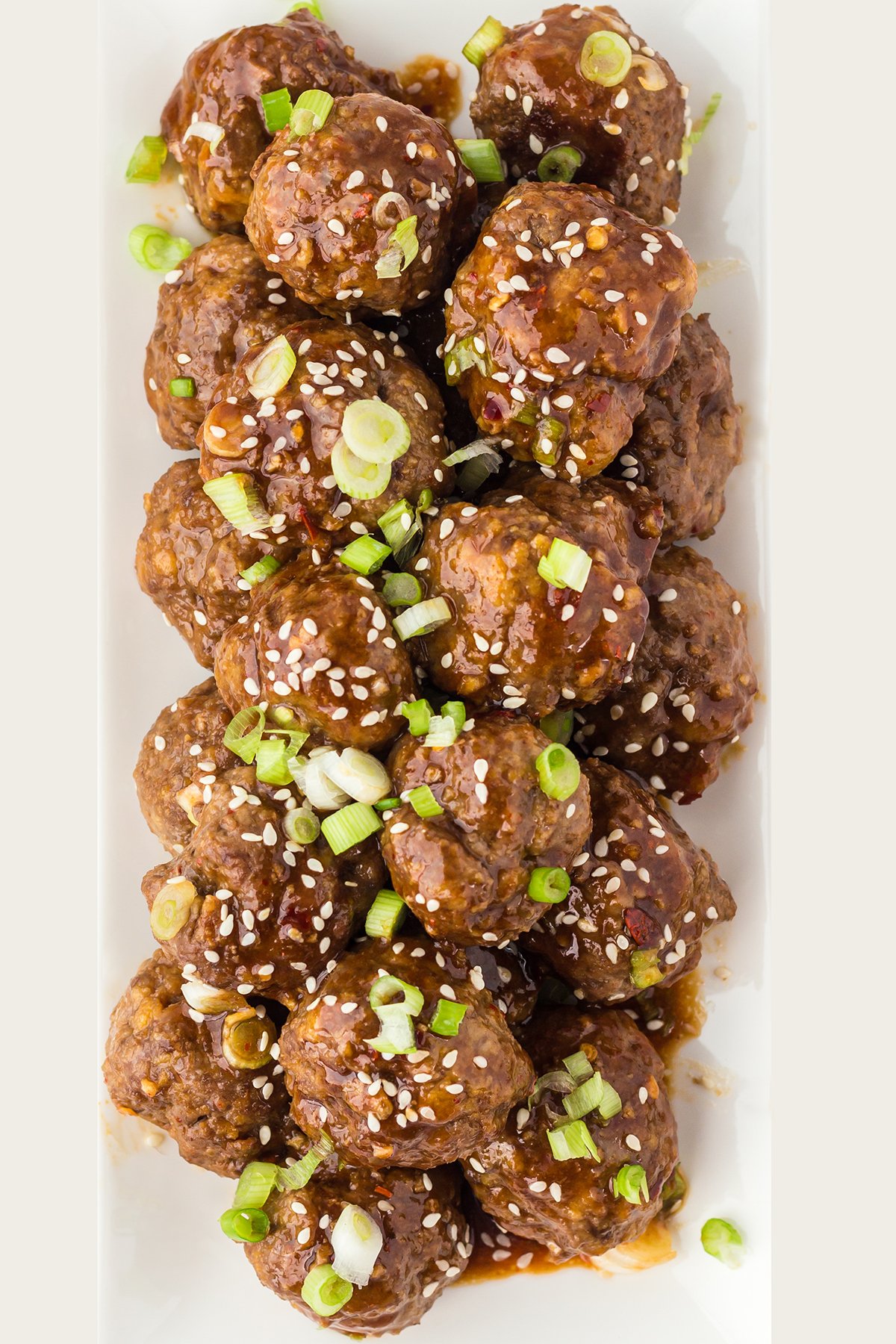 A white rectangular serving platter filled with saucy Asian meatballs garnished with green onions and sesame seeds.