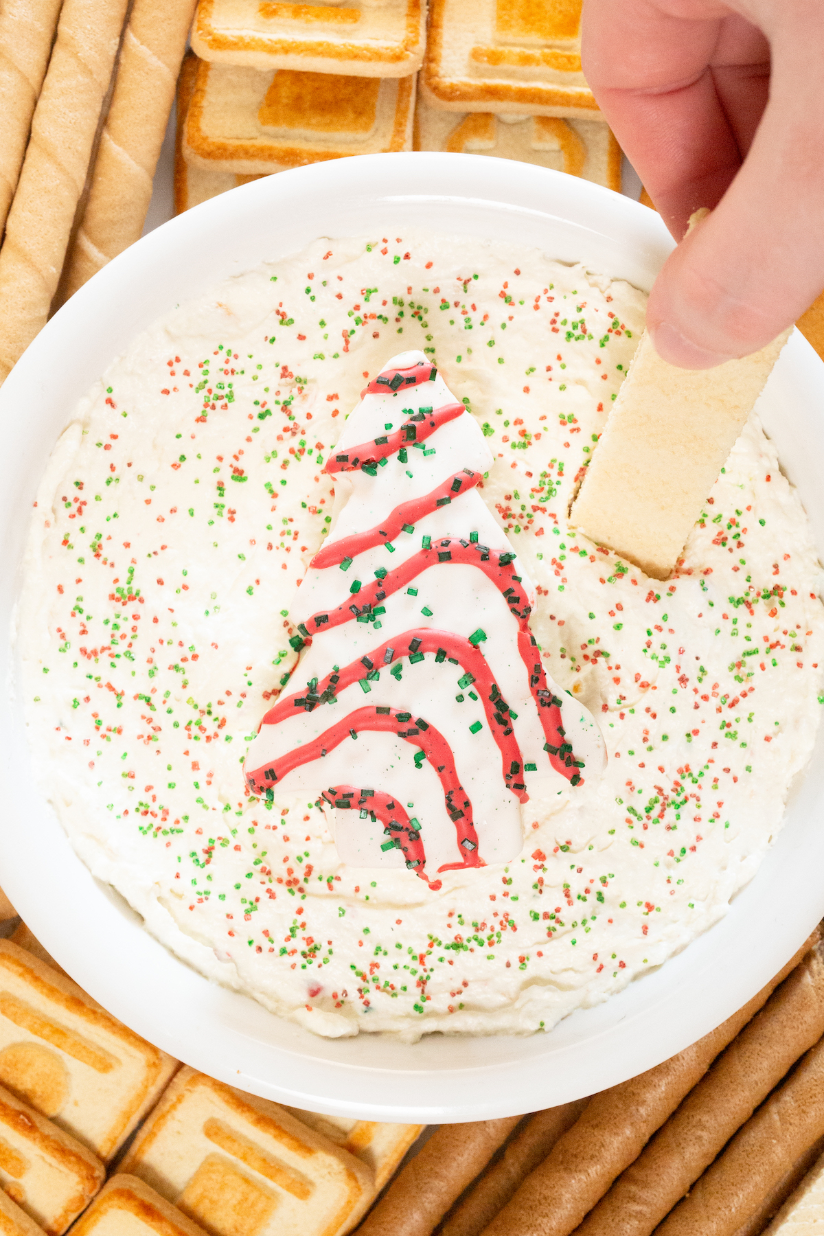 A hand dips a vanilla sugar wafer into a bowl of Christmas Tree Cake Dip.