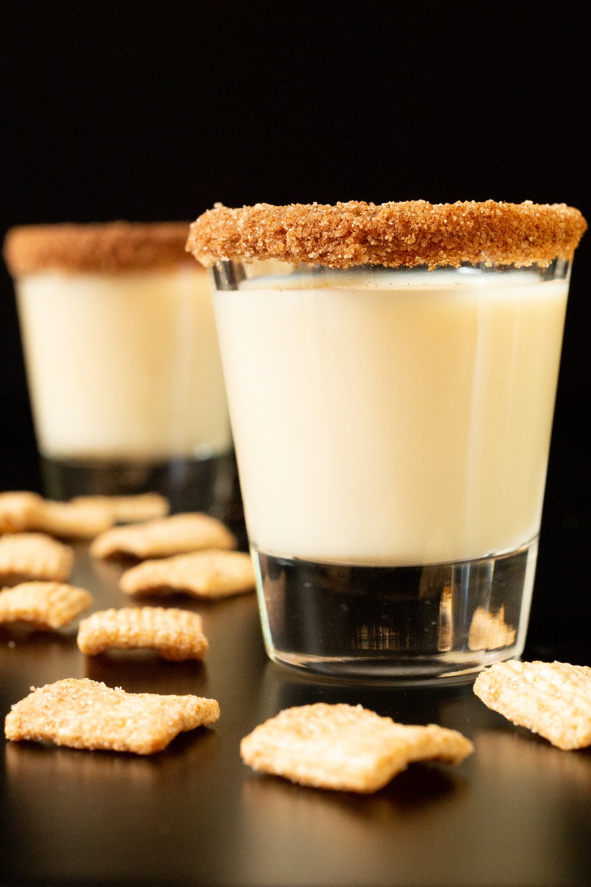 Two shot glasses each rimmed with cinnamon and sugar and filled with a Cinnamon Toast Crunch shot. Pieces of the cereal surround them.