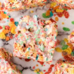 Cut squares of fruity pebble treats on a marble board with loose pieces of the cereal.