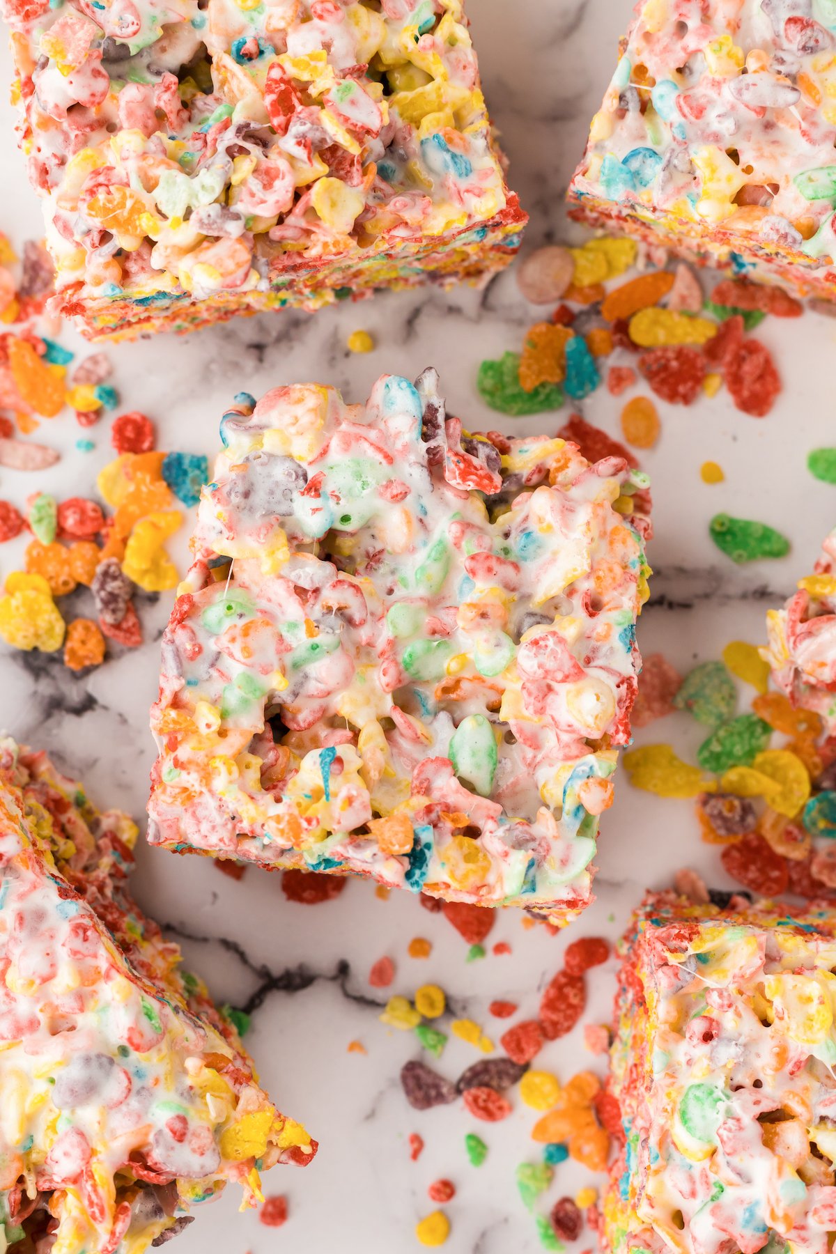 Cut squares of fruity pebble treats on a marble board with loose pieces of the cereal.