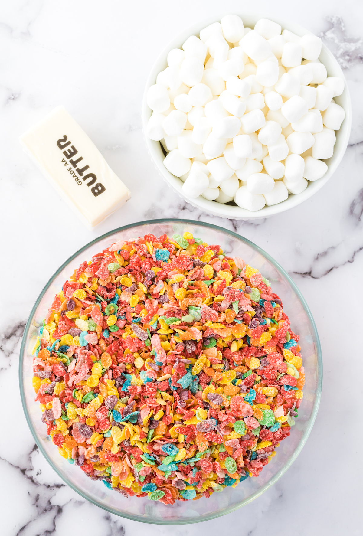 Two prep bowls one filled with fruity pebbles and the other mini marshmallows sitting next to a stick of butter.