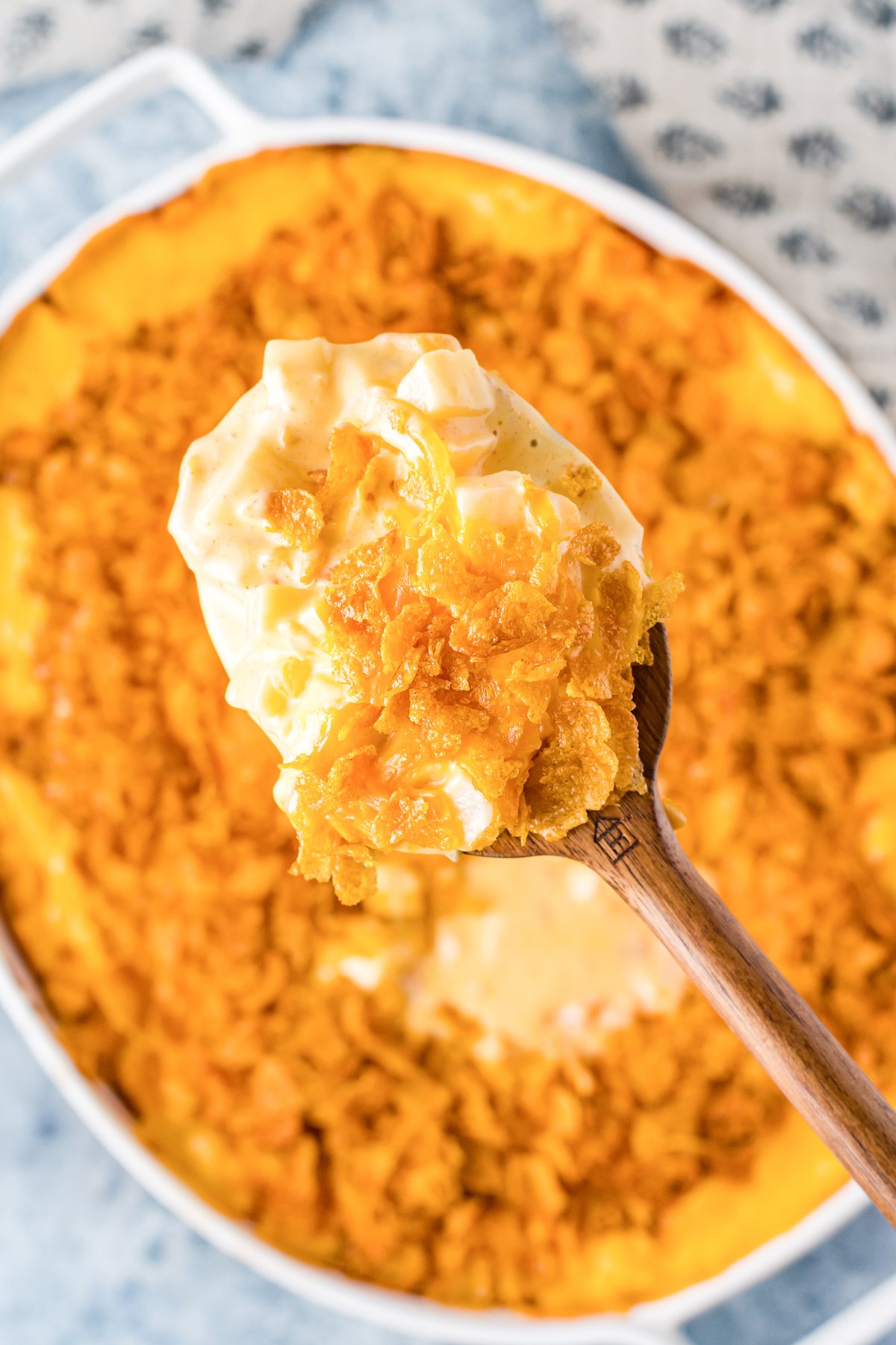 A wooden spoon is filled with funeral potatoes. The casserole dish is out of focus in the background.